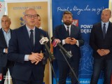 Mężczyzna w okularach przemawia do mediów, a za nim grupa przedstawicieli władz lokalnych
