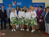 Uczestniczki w kwiecistych spódnicach i białych koszulach otrzymują wyróżnienie, robiąc sobie pamiątkowe zdjęcie z organizatorami